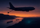 Editorial image of a gray aerial refueling tanker above a desert coastline and narrow shipping strait at dusk, with distant oil tankers and radar-like light below.